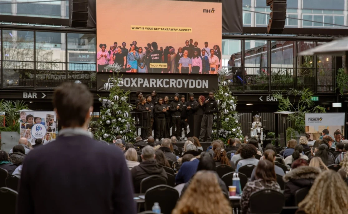 Young people on stage at a Boxpark Croydon event, presenting to a large seated audience. A screen behind them displays "What is your key takeaway advice?" under the Reaching Higher brand.