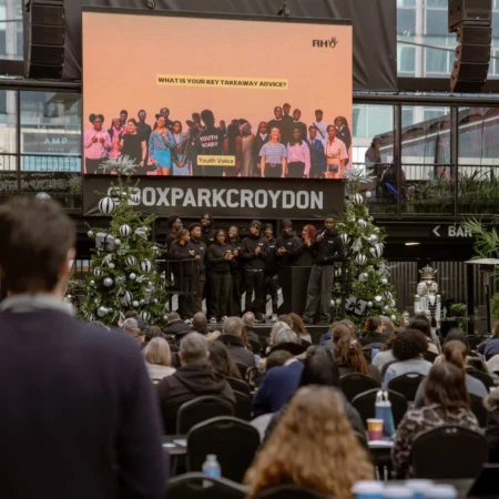 Young people on stage at a Boxpark Croydon event, presenting to a large seated audience. A screen behind them displays "What is your key takeaway advice?" under the Reaching Higher brand.