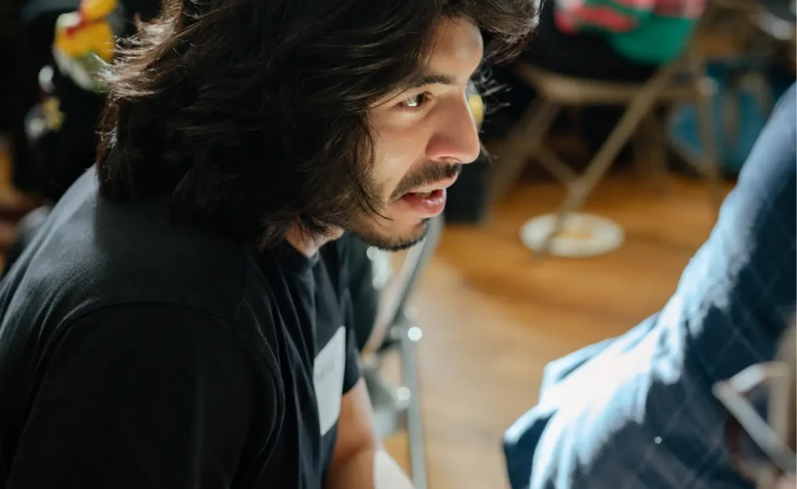Person with long dark hair speaking during a collaborative workshop session, seated in a light-filled event space.