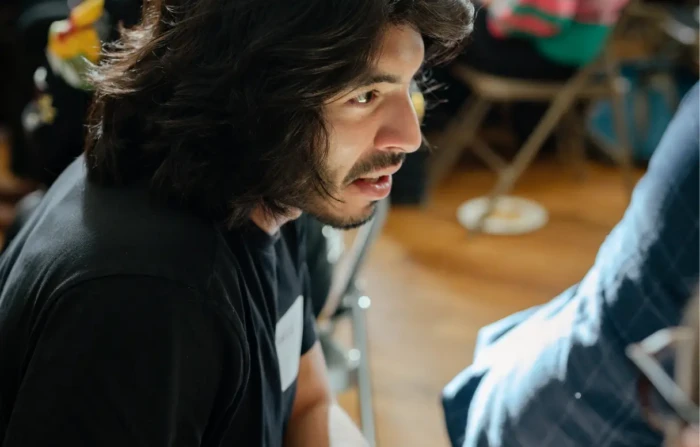 Person with long dark hair speaking during a collaborative workshop session, seated in a light-filled event space.