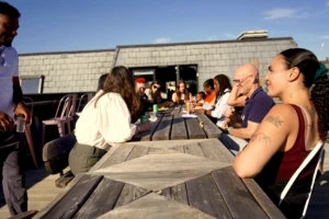 A diverse group of people sit around a large outdoor wooden picnic table on a sunny rooftop terrace, talking and laughing together at a Camden Giving community event.